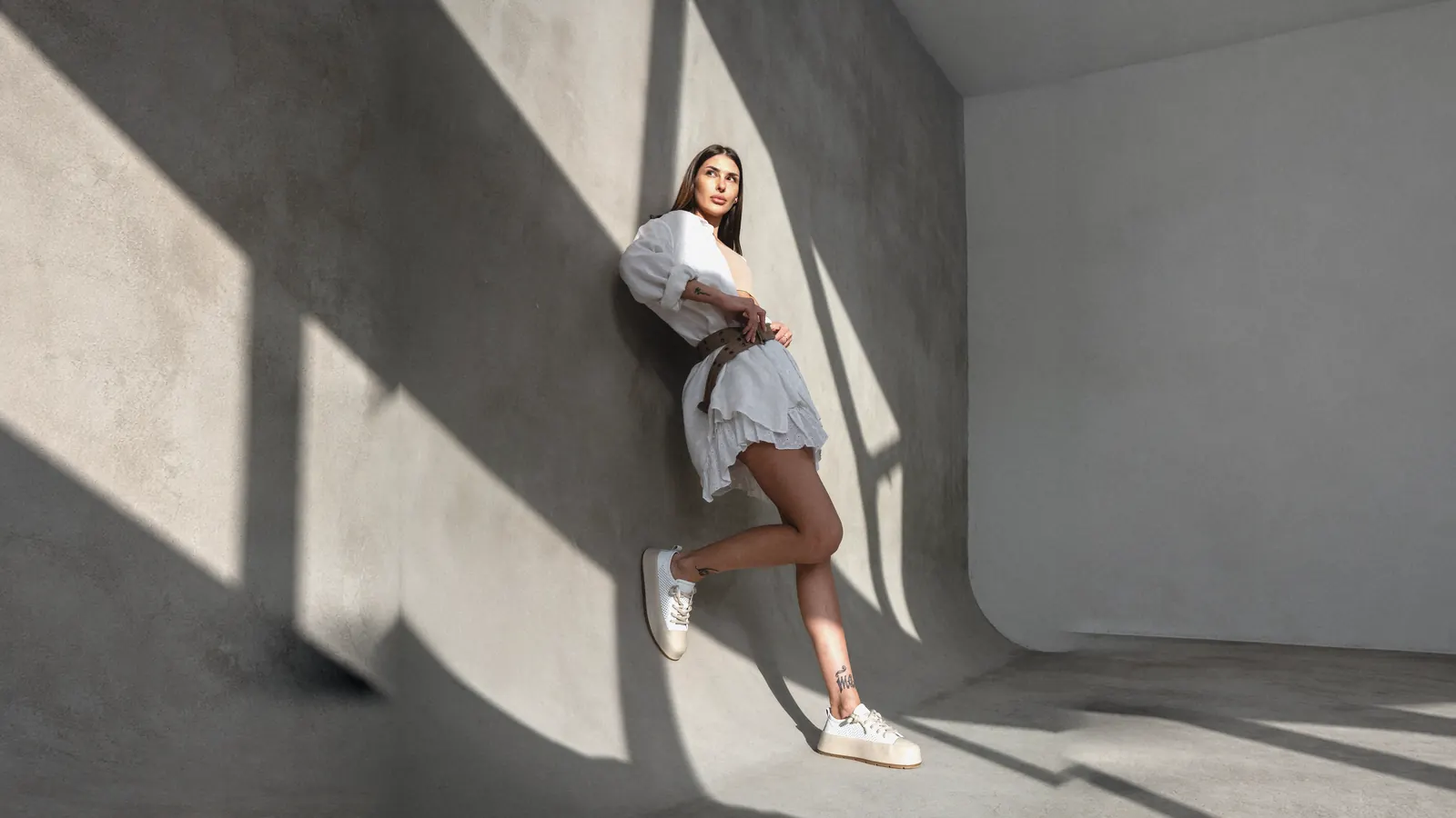 A woman in white Babylon sneakers against a sunlit concrete wall