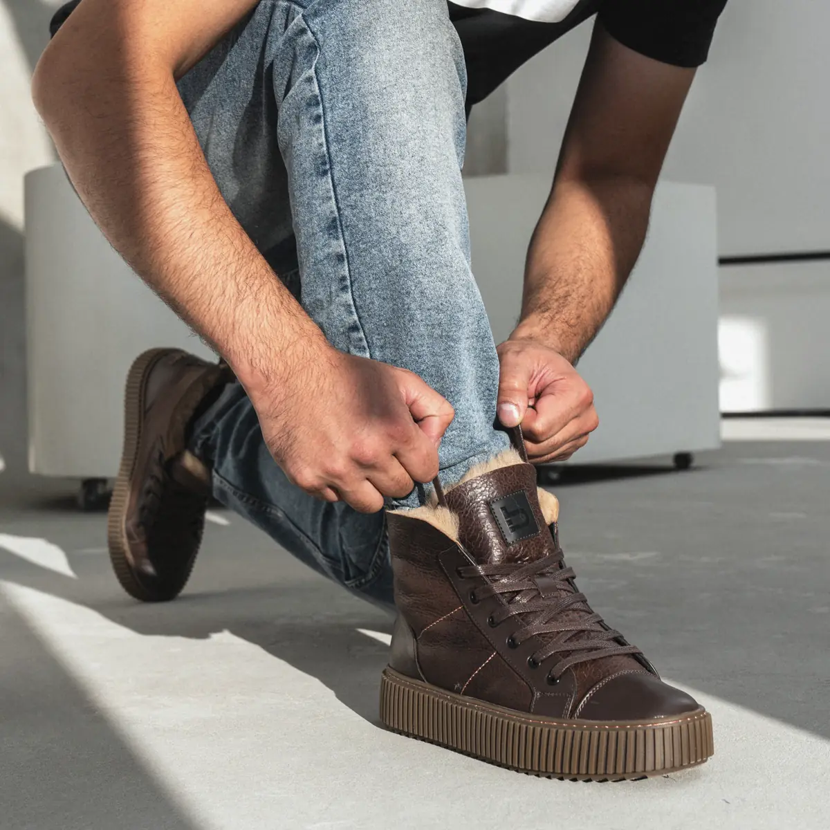 Man in light jeans lacing dark brown leather Frost-84 sheepskin boots with a fleece collar, the other boot behind