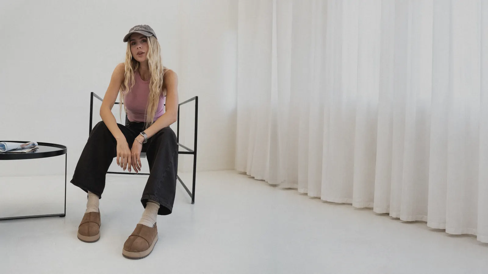 A woman in a cap and black jeans sits on a chair in a bright room wearing tan nubuck Babylon MARY-94 sheepskin slip-ons and white socks