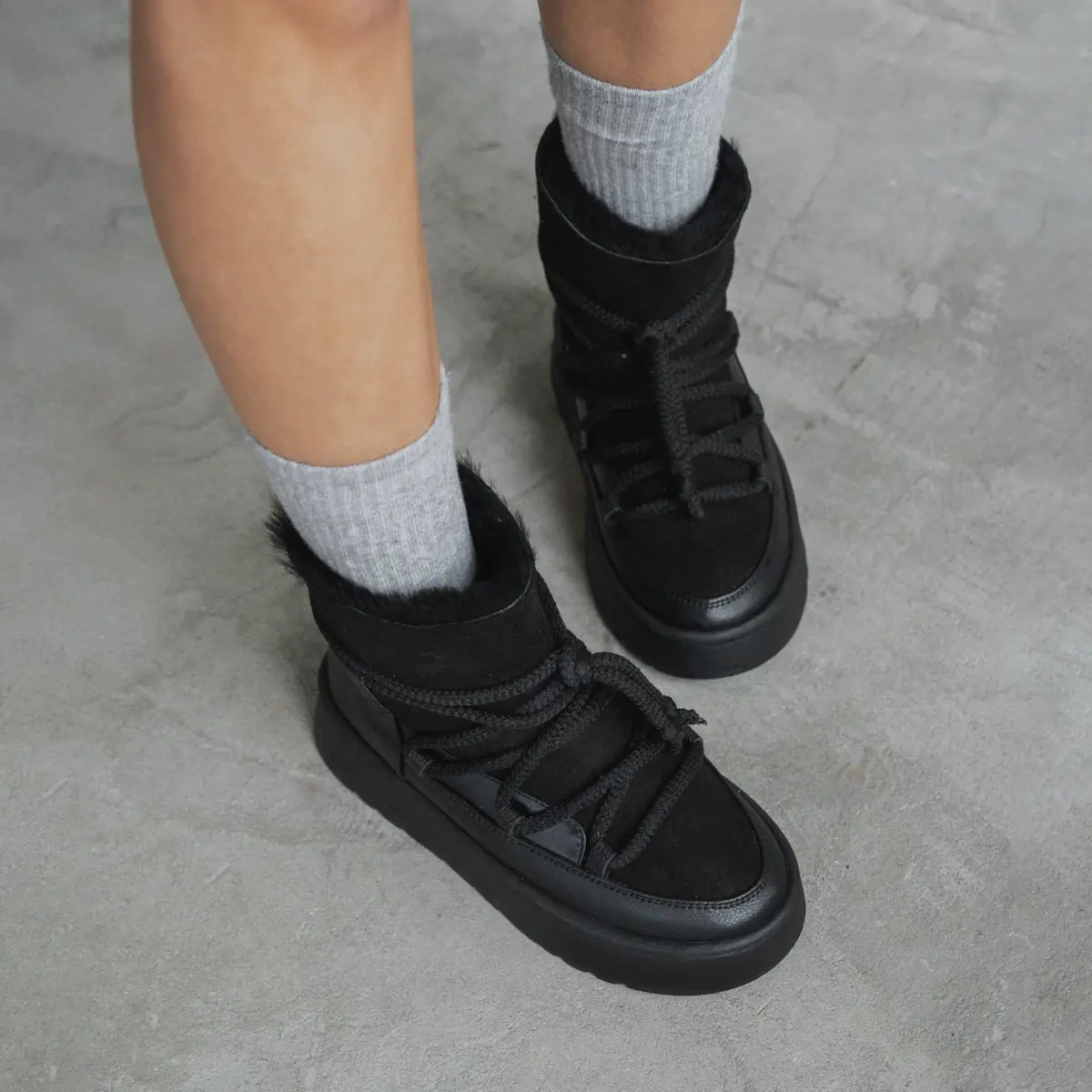 Feet in grey socks wearing black Babylon UG-1-41 women's sheepskin boots with a fur collar and braided laces on a concrete floor
