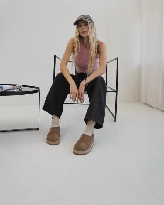 A woman in a cap and black jeans sits on a chair in a bright room wearing tan nubuck Babylon MARY-94 sheepskin slip-ons and white socks
