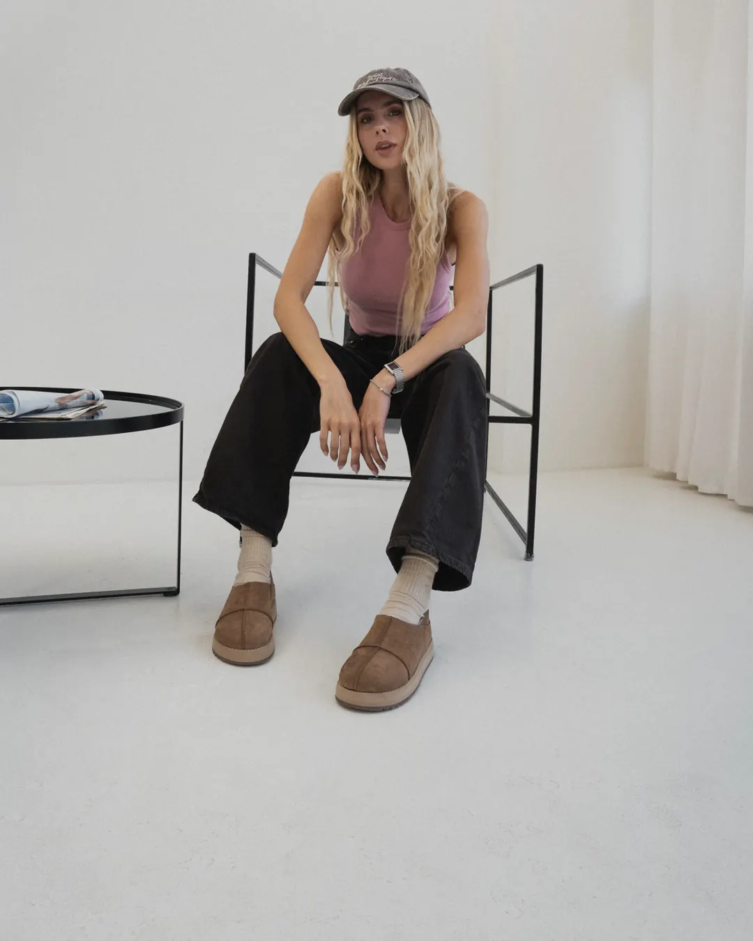 A woman in a cap and black jeans sits on a chair in a bright room wearing tan nubuck Babylon MARY-94 sheepskin slip-ons and white socks