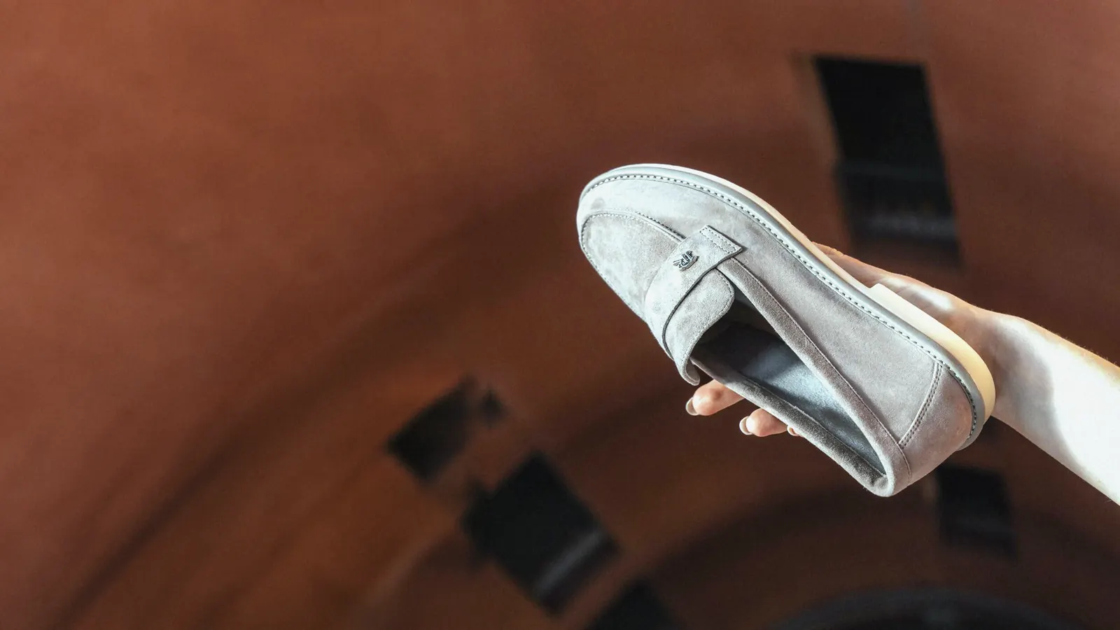 A Babylon suede loafer held in a hand against a terracotta background