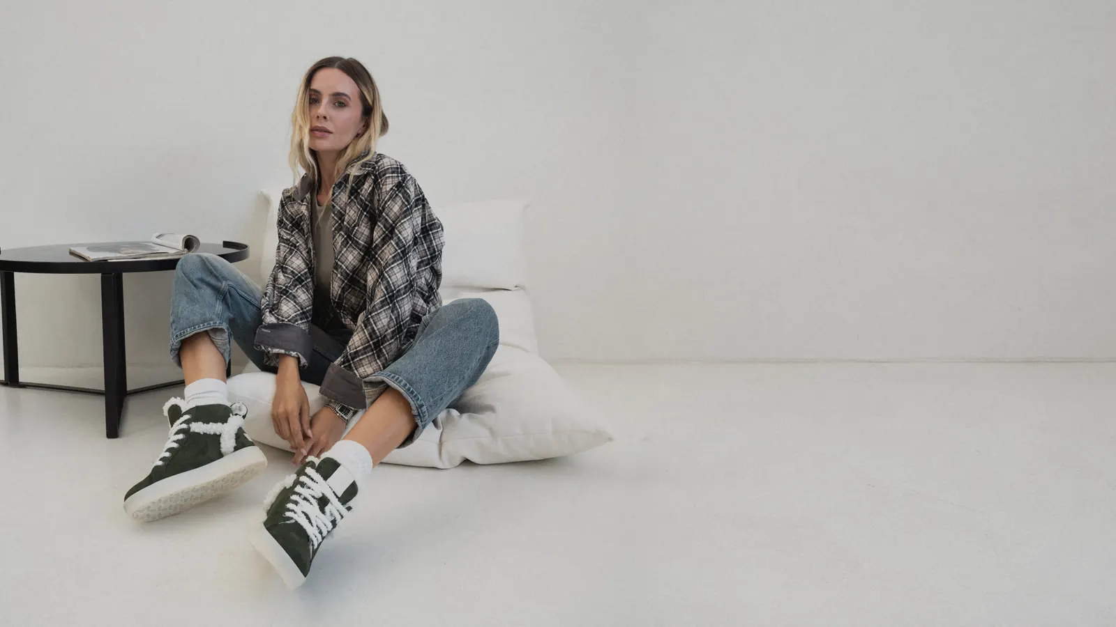 A woman in a check shirt and jeans sits on a cushion wearing green fur-lined Babylon Glori-18 nubuck boots