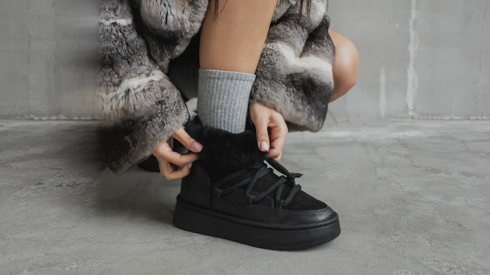 A woman in a fur coat and grey socks laces up black Babylon UG-1-41 sheepskin boots