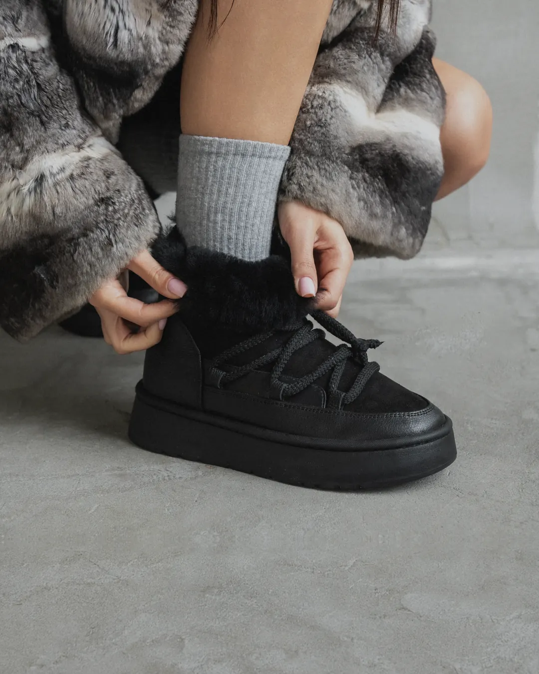 A woman in a fur coat and grey socks laces up black Babylon UG-1-41 sheepskin boots