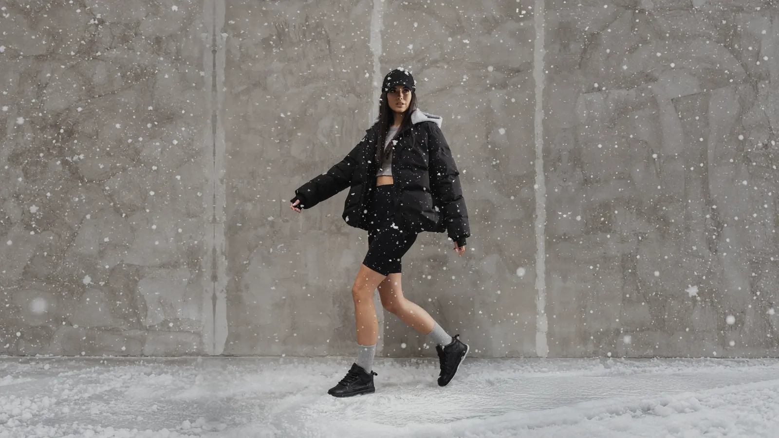 A woman in a black puffer jacket walks through snow in black insulated Babylon Space-81-X sneakers and grey socks