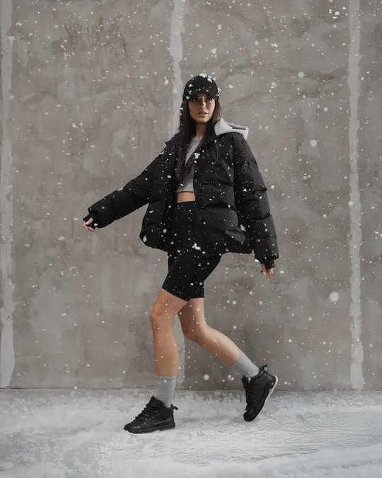 A woman in a black puffer jacket walks through snow in black insulated Babylon Space-81-X sneakers and grey socks